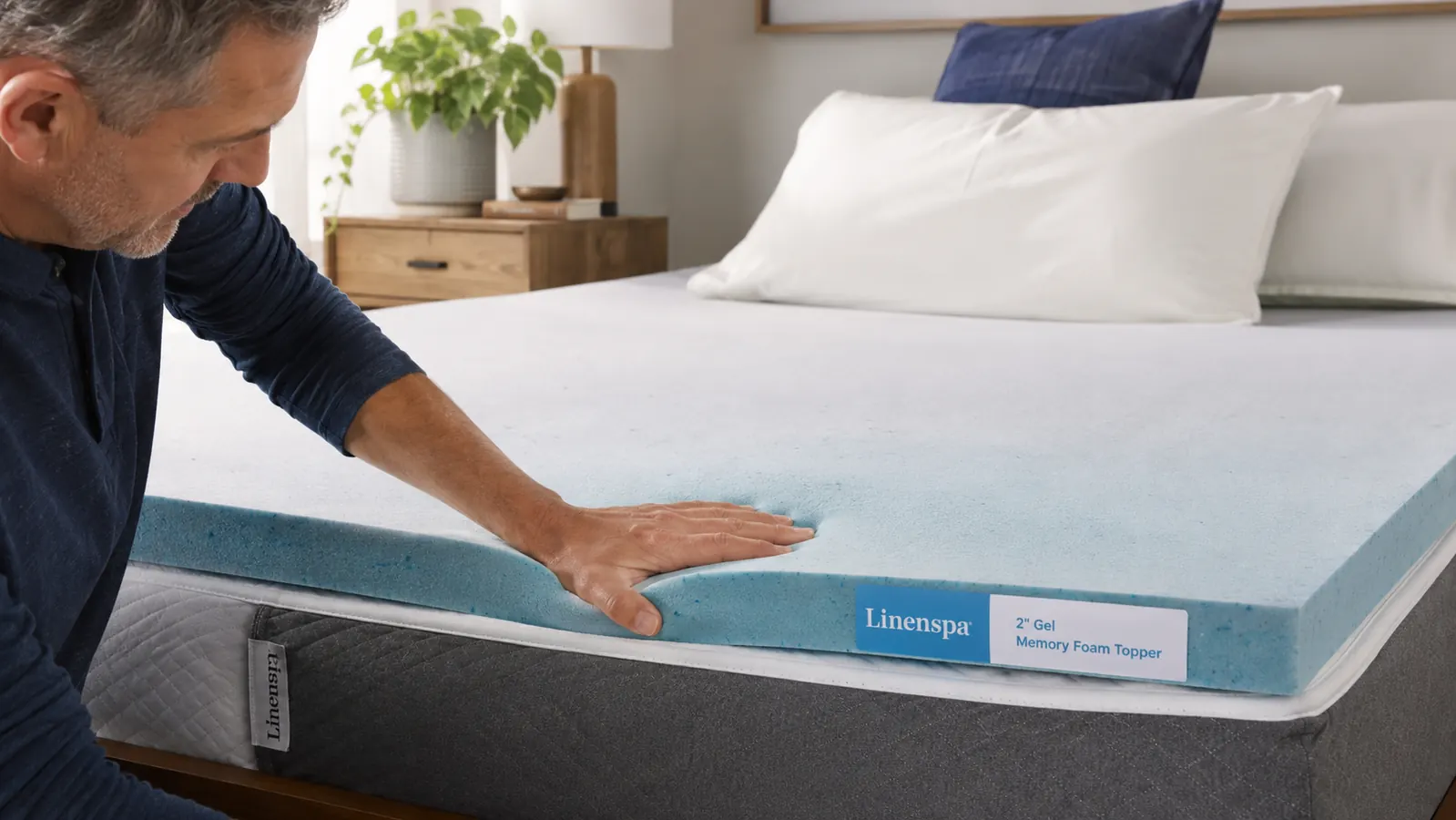 A hand pressing into a white gel-infused memory foam mattress topper on top of a bed, showing the foam slowly rebounding