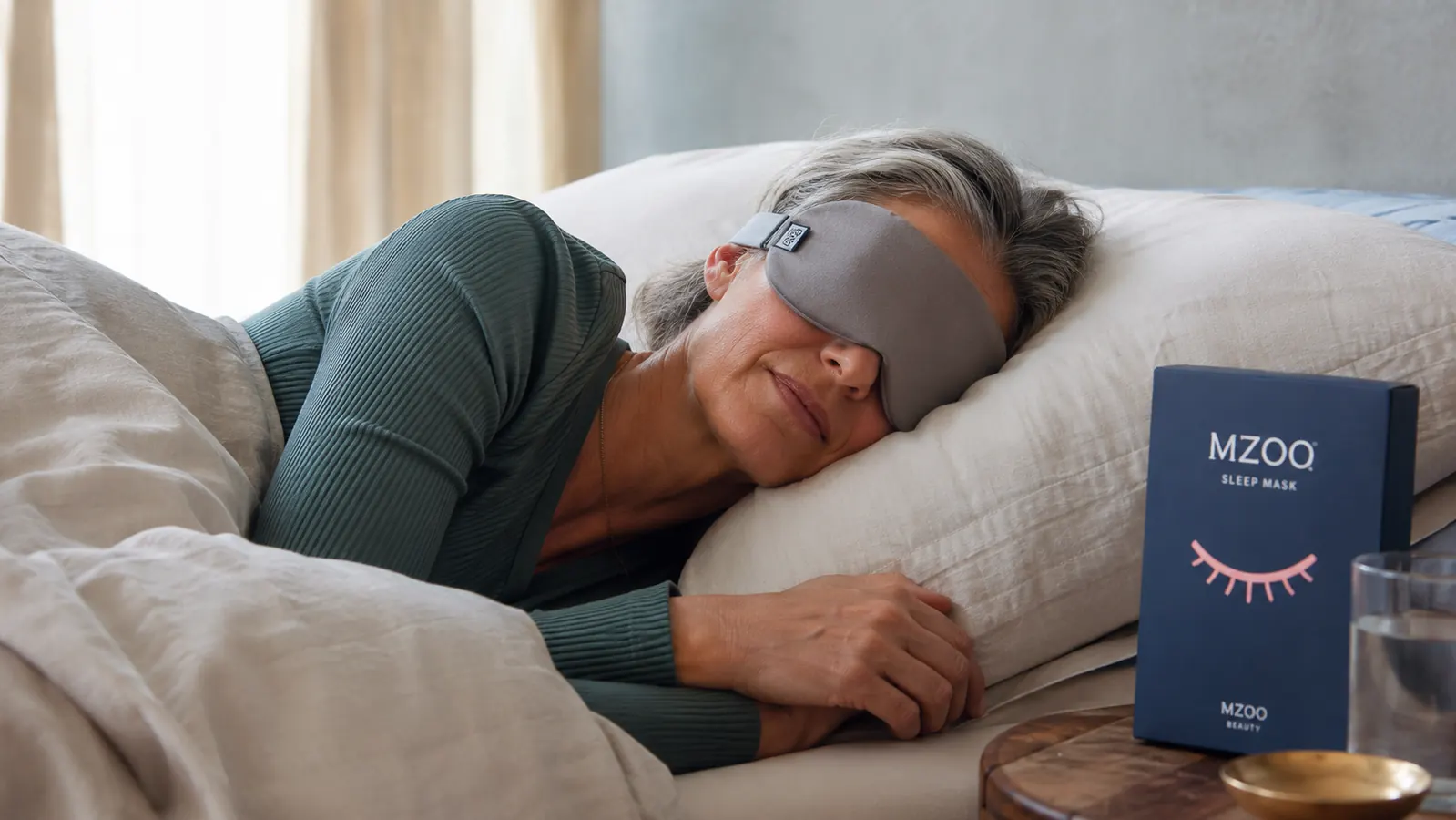 Person lying on their side wearing the MZOO sleep mask with the strap visible, faint mark under one eye from an earlier ill-fitted night