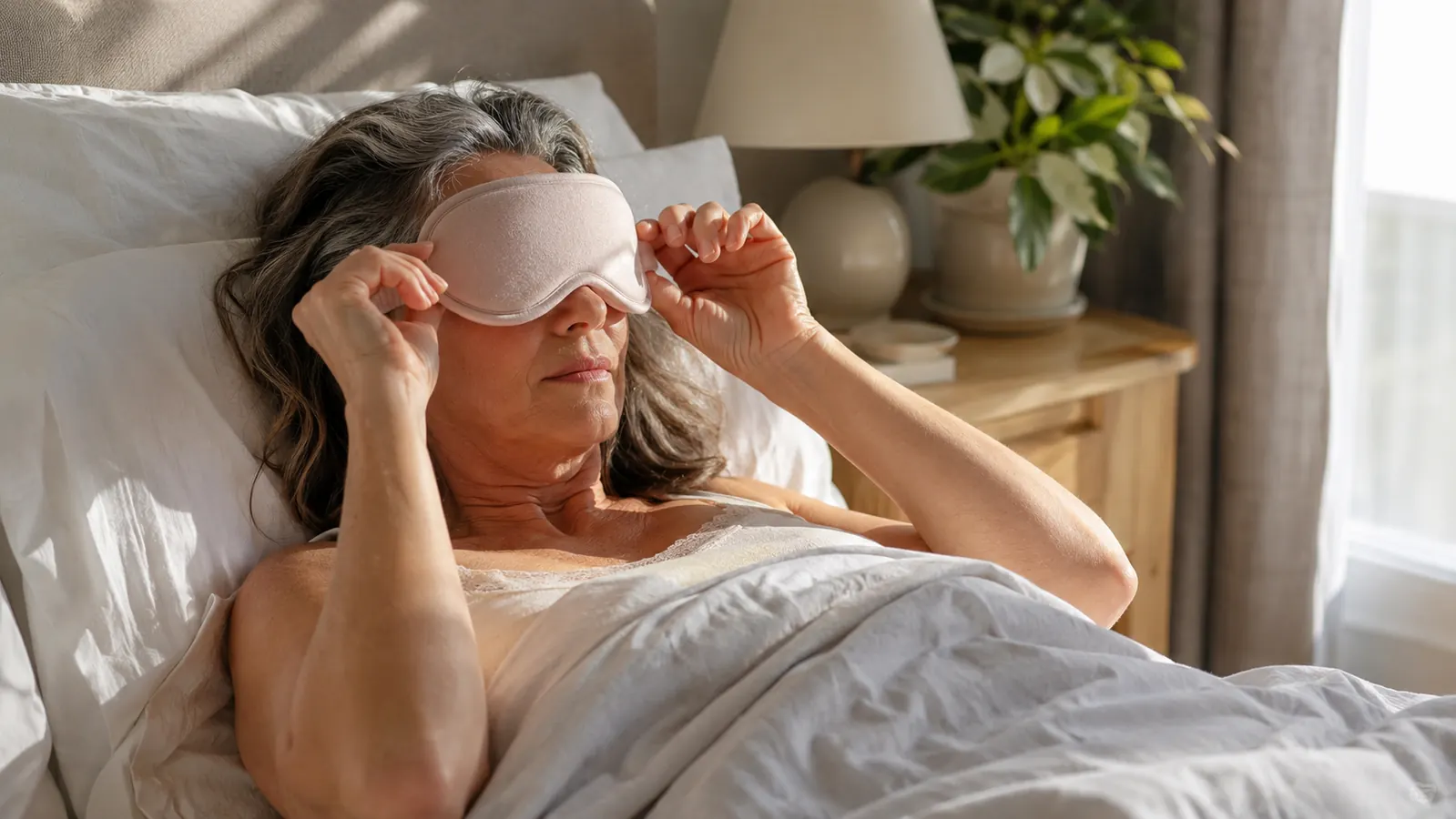 Person lying down in a bright bedroom pulling a contoured sleep mask down over their eyes during the day